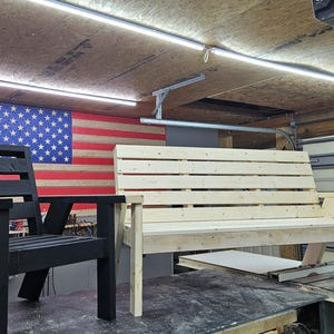 May include: Two wooden benches, one black and one natural wood, are shown in a workshop setting. The black bench has a slatted back and seat, while the natural wood bench has a slatted back and seat and a wide armrest on one side. Both benches are unfinished.