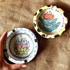 以下が含まれることがあります： 手描きのデザインが施された2つのセラミック灰皿。1つは炎のあるさくらんぼ、もう1つは花が特徴です。灰皿は、黄色、黒、青、ピンクなどのカラフルなアクセントが付いた白いベースです。灰皿は丸く、小さな切り込みがあります。