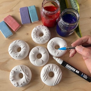May include: White ceramic donut ornaments with different patterns. Jars of red and blue paint, plus small rectangular ceramic pieces in pastel shades, are arranged on a wooden surface. A blue marker is also present.