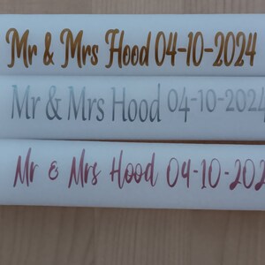 May include: Three white cylindrical objects with the text "Mr & Mrs Hood 04-10-2024" in gold, silver, and pink lettering.