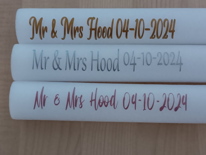 May include: Three white cylindrical objects with the text "Mr & Mrs Hood 04-10-2024" in gold, silver, and pink lettering.