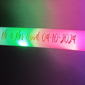 May include: A white light-up stick with a green, pink, and purple glow. The stick has the text "Mr & Mrs Hood 04-10-2024" written on it.