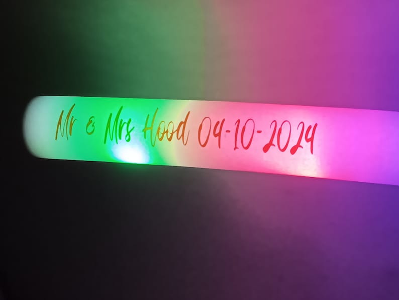 May include: A white light-up stick with a green, pink, and purple glow. The stick has the text "Mr & Mrs Hood 04-10-2024" written on it.