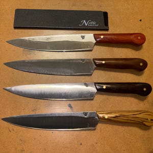 May include: Four kitchen knives with silver-toned blades and wooden handles in varying shades of red, brown, dark brown, and light brown with grain patterns. Each handle features two brass rivets. A black sheath with the text Noble home & chef is included.