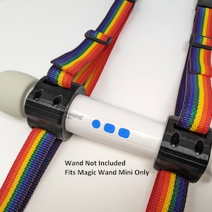 Puede incluir: Un arnés blanco y negro con correas de arcoíris para un Magic Wand Mini. El arnés tiene dos soportes de plástico negro con agujeros para sujetar las correas. El texto "Wand Not Included" y "Fits Magic Wand Mini Only" está impreso debajo del arnés.