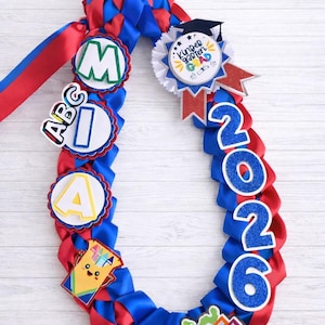 May include: A graduation lei in red and blue ribbon, adorned with decorative elements. Features the letters "M", "A", "I", and "A", along with the year "2026" and a badge that says "Kindergarten Grad".
