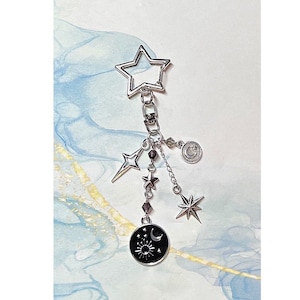 May include: Silver star-shaped keychain with dangling charms, including a black circle with a moon and sun design, a star, and a crescent moon charm.