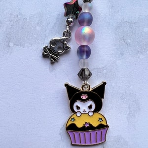 Pode incluir: Um amuleto de cupcake preto e branco com um nariz rosa e cobertura amarela. O amuleto est&aacute; preso a uma corrente prateada com contas iridescentes e um amuleto de caveira prateada.