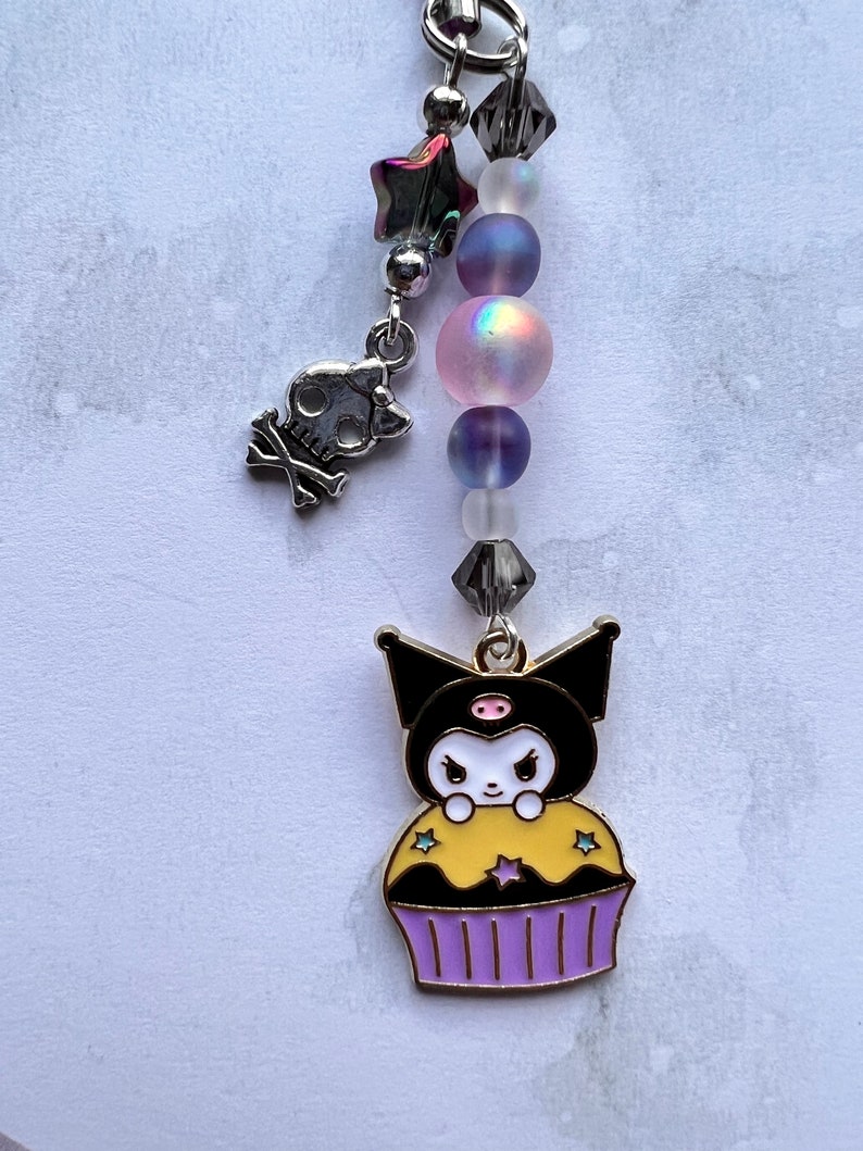 Pode incluir: Um amuleto de cupcake preto e branco com um nariz rosa e cobertura amarela. O amuleto est&aacute; preso a uma corrente prateada com contas iridescentes e um amuleto de caveira prateada.