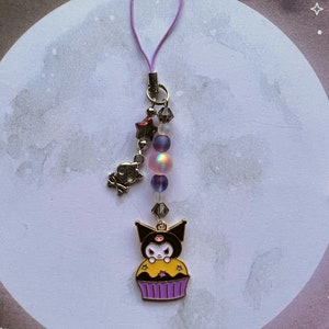 Pode incluir: Um amuleto de cupcake preto e amarelo com um personagem de gato preto no topo. O amuleto est&aacute; preso a um cord&atilde;o roxo com contas iridescentes e um amuleto de estrela prateada.