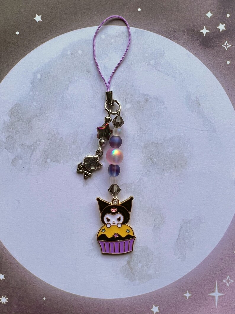 Pode incluir: Um amuleto de cupcake preto e amarelo com um personagem de gato preto no topo. O amuleto est&aacute; preso a um cord&atilde;o roxo com contas iridescentes e um amuleto de estrela prateada.