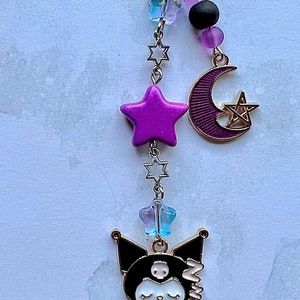 Pode incluir: Um amuleto de gato de desenho animado preto e branco com uma estrela roxa, uma lua crescente com um pentagrama e contas de estrelas azuis e roxas em uma corrente dourada.
