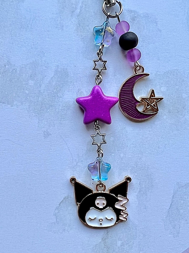 Pode incluir: Um amuleto de gato de desenho animado preto e branco com uma estrela roxa, uma lua crescente com um pentagrama e contas de estrelas azuis e roxas em uma corrente dourada.