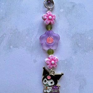 Pode incluir: Um amuleto de personagem de desenho animado preto e branco com um nariz rosa e um amuleto de flor roxa. O amuleto est&aacute; preso a um cord&atilde;o roxo com um fecho prateado e dois amuletos de flores rosa e brancas.