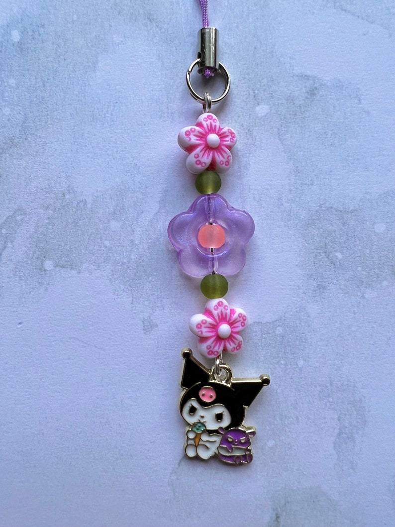 Pode incluir: Um amuleto de personagem de desenho animado preto e branco com um nariz rosa e um amuleto de flor roxa. O amuleto est&aacute; preso a um cord&atilde;o roxo com um fecho prateado e dois amuletos de flores rosa e brancas.