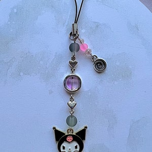 Pode incluir: Um amuleto de personagem preto e branco com um donut, preso a um cord&atilde;o preto com contas prateadas, uma conta roxa e um amuleto de cora&ccedil;&atilde;o prateado.