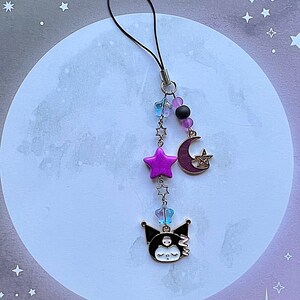 Pode incluir: Um acess&oacute;rio de telefone com um personagem de desenho animado preto e branco, uma estrela rosa, uma lua crescente e contas roxas em um cord&atilde;o preto.