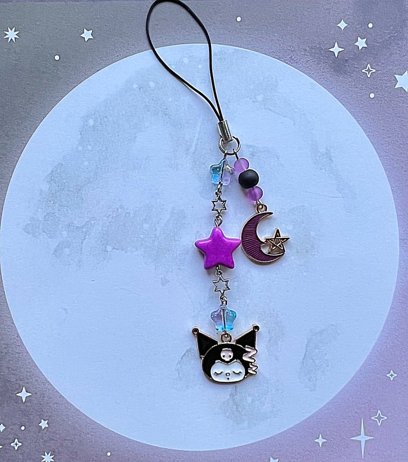 Pode incluir: Um acess&oacute;rio de telefone com um personagem de desenho animado preto e branco, uma estrela rosa, uma lua crescente e contas roxas em um cord&atilde;o preto.