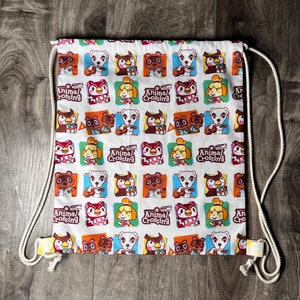 Könnte beinhalten: Weiße Kordeltasche mit einem sich wiederholenden Muster von Animal Crossing-Charakteren. Zu den Charakteren gehören Dorfbewohner wie Tom Nook, Isabelle und K.K. Slider. Die Tasche trägt den Text "Animal Crossing", der sich im gesamten Muster wiederholt.