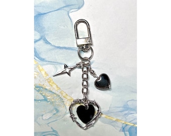 May include: Silver-coloured keychain with a lobster clasp, featuring a star charm, a small heart, and a heart-shaped pendant with a black centre, wrapped in a wire design. Ideal for keys or bags.