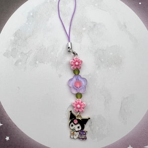 Pode incluir: Um amuleto de personagem de desenho animado preto e branco com um amuleto de flor rosa e contas de flores roxas e verdes em um cord&atilde;o roxo.