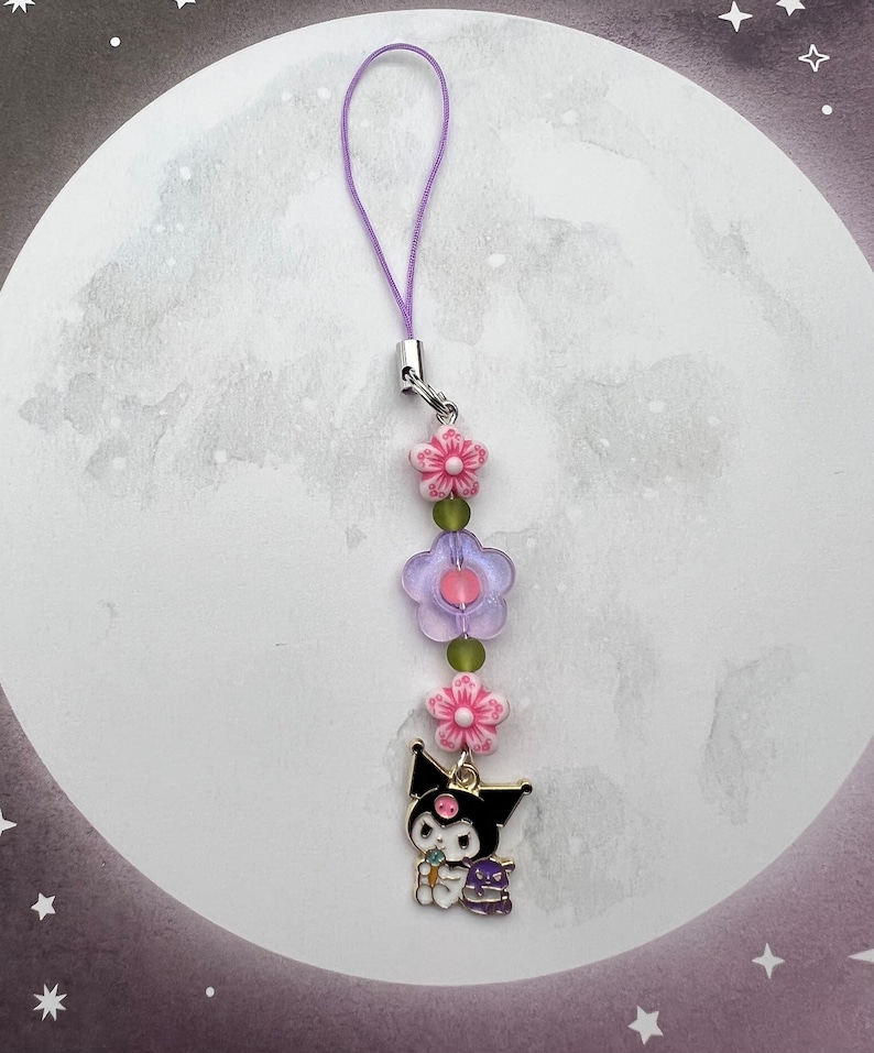 Pode incluir: Um amuleto de personagem de desenho animado preto e branco com um amuleto de flor rosa e contas de flores roxas e verdes em um cord&atilde;o roxo.