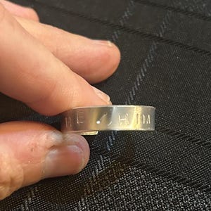 May include: A silver ring with the words "HE/HIM" engraved on the outside. The ring is held between two fingers against a dark background.