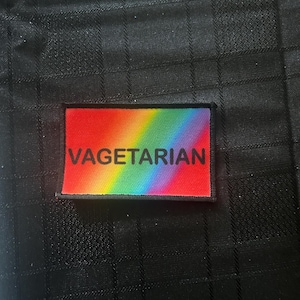 Könnte beinhalten: Ein rechteckiger Aufnäher mit dem Wort "VAGETARIAN" in weißen Großbuchstaben auf rotem Hintergrund. Der Aufnäher hat ein Regenbogen-Verlaufsdesign. Der Aufnäher befindet sich auf einem schwarzen, strukturierten Stoff.
