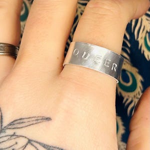Peut inclure: Une bague en argent avec le mot "QUEER" gravé dessus.