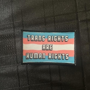 以下が含まれることがあります： 白い大文字で「TRANS RIGHTS ARE HUMAN RIGHTS」と書かれた長方形のパッチ。パッチは、水色、ピンク、白のトランスジェンダープライドフラッグの色を特徴としています。パッチは黒のテクスチャード加工された背景に設定されています。