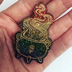 May include: A rainbow glitter cauldron with gold accents and a swirling smoke design. The cauldron is a decorative item with a whimsical and magical theme.