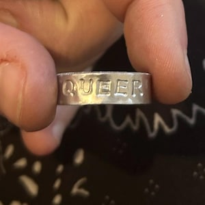 May include: A silver ring with the word "QUEER" embossed on it.