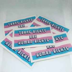 May include: A stack of white stickers with the text "Trans Rights Are Human Rights" in blue and pink letters. The stickers feature a blue, pink, and white colour scheme reminiscent of the transgender pride flag.