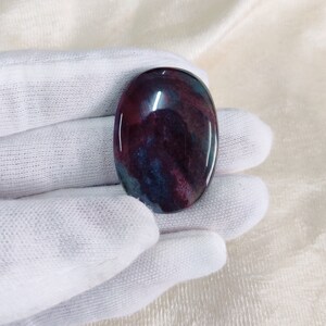Peut inclure: Une pierre polie lisse de forme ovale avec un motif tourbillonnant de teintes rouge foncé, violet et bleu.