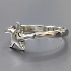 Könnte beinhalten: Ein silberner Ring mit einem Seestern-Design. Der Ring hat eine polierte Oberfläche, wobei sich der Seestern vom Band erstreckt. Der Ring ist vor einem grauen Hintergrund platziert, der die Details des Schmucks hervorhebt.