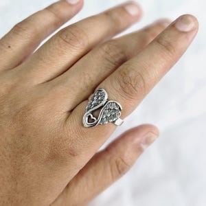May include: A silver-colored ring featuring a design of angel wings and a heart-shaped cutout. The ring is worn on a finger, showcasing the intricate details of the wing feathers and the heart. The ring is a piece of jewelry.
