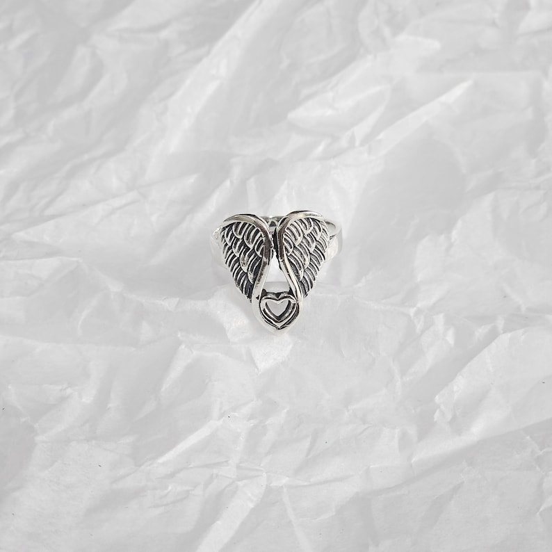 May include: A silver-colored ring featuring a detailed wing design with a heart-shaped cutout at the base. The ring is set against a white, textured background, highlighting its intricate details and metallic sheen.