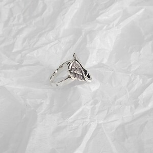 May include: A silver-colored ring featuring a detailed wing design. The ring has a polished finish and a band with a subtle texture. The wing design is the focal point, with intricate feather detailing. The ring is likely made of sterling silver.
