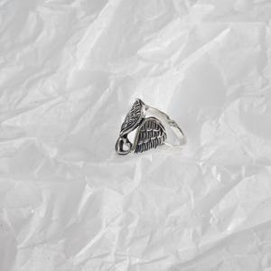 May include: A silver-colored ring featuring an angel wing design. The ring has detailed feather textures and a heart-shaped accent. The ring is set against a white, textured background.