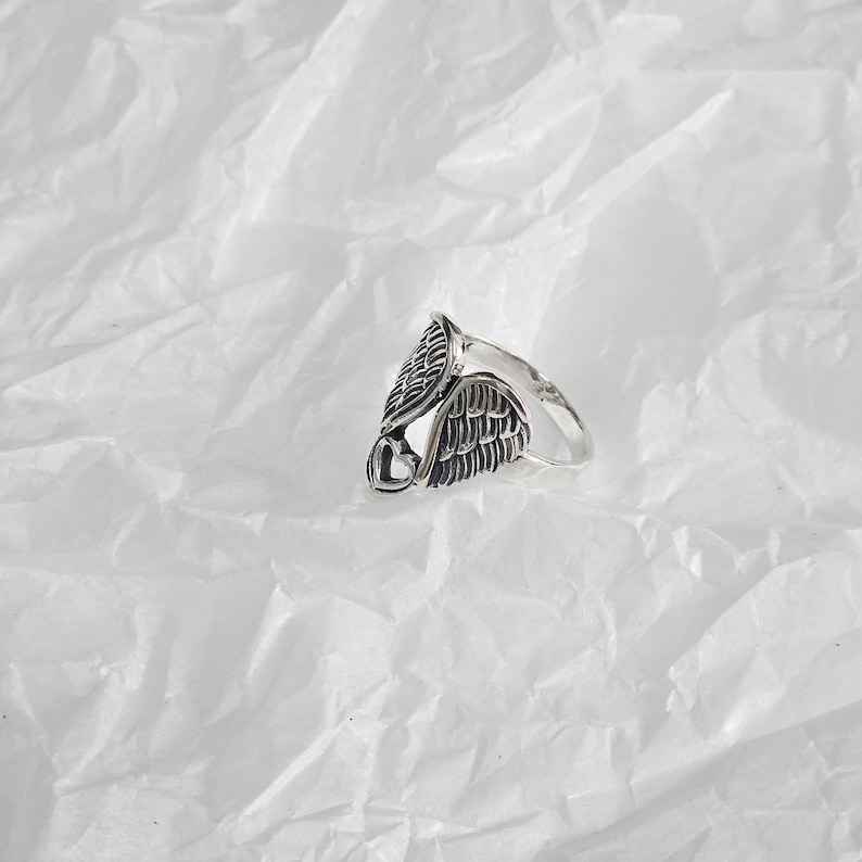 May include: A silver-colored ring featuring an angel wing design. The ring has detailed feather textures and a heart-shaped accent. The ring is set against a white, textured background.