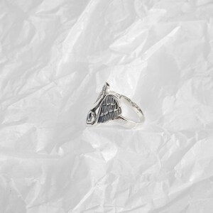 May include: A silver-toned ring featuring a detailed wing design. The ring has a textured finish and a prominent wing motif, with intricate feather detailing. The ring is set against a white, textured background, highlighting its metallic sheen.