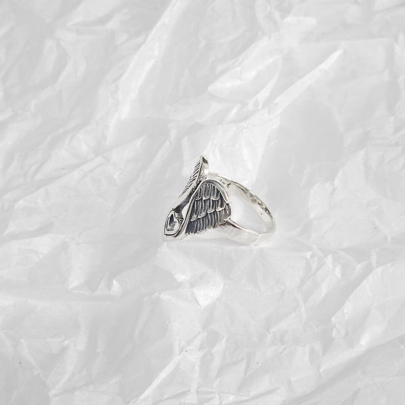 May include: A silver-toned ring featuring a detailed wing design. The ring has a textured finish and a prominent wing motif, with intricate feather detailing. The ring is set against a white, textured background, highlighting its metallic sheen.