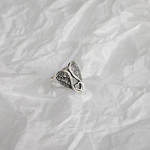 May include: A silver-toned ring featuring a wing design with a heart-shaped cutout. The ring has detailed feather textures and is set against a white, textured background. The ring's design is symmetrical and detailed.