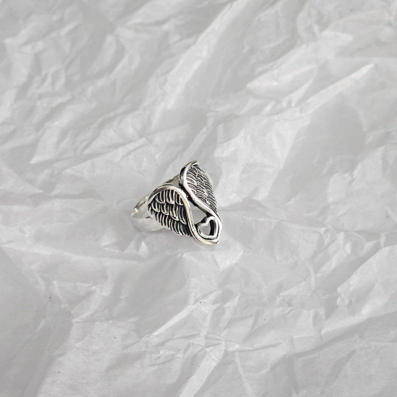 May include: A silver-toned ring featuring a wing design with a heart-shaped cutout. The ring has detailed feather textures and is set against a white, textured background. The ring's design is symmetrical and detailed.