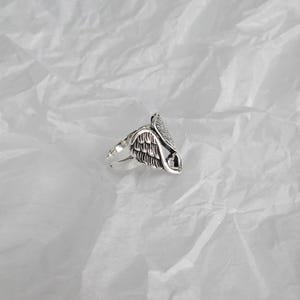 May include: A silver ring featuring a detailed wing design with a heart-shaped cutout. The ring has a polished finish and is set against a textured white background. The ring's design is intricate and detailed.