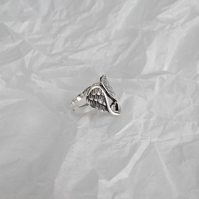 May include: A silver ring featuring a detailed wing design with a heart-shaped cutout. The ring has a polished finish and is set against a textured white background. The ring's design is intricate and detailed.