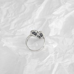 May include: A silver ring with a floral design. The ring features a detailed, openwork flower motif with dark accents. The band is thin and polished, and the ring is set against a white, textured background.