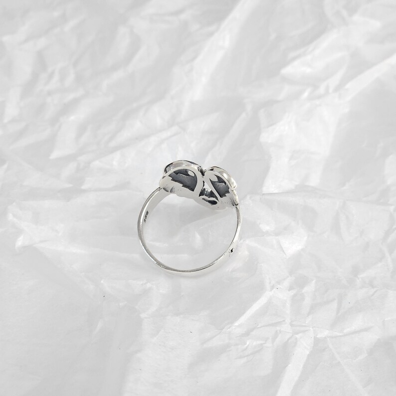 May include: A silver ring with a floral design. The ring features a detailed, openwork flower motif with dark accents. The band is thin and polished, and the ring is set against a white, textured background.