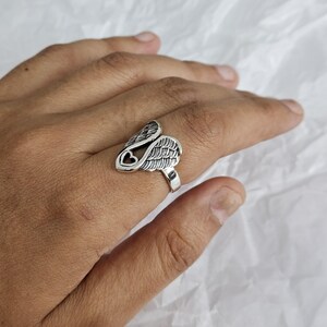 May include: A silver-colored ring featuring a wing design with a heart-shaped cutout. The ring has detailed feather textures and is worn on a finger. The ring is made of a shiny metal.