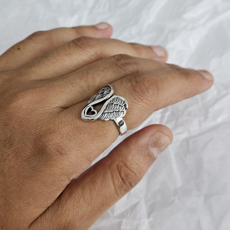 May include: A silver-colored ring featuring a wing design with a heart-shaped cutout. The ring has detailed feather textures and is worn on a finger. The ring is made of a shiny metal.